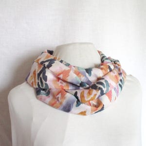 Op de afbeelding: Een kleurrijke infinity sjaal met een aquarel bloemenpatroon. De sjaal heeft een mix van oranje, roze, paarse en groene abstracte vormen op een witte achtergrond. De sjaal is om een mannequin gewikkeld en toont het ontwerp en de stijl.