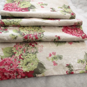 Op de afbeelding: Een beige linnen tafelloper met een bloemenpatroon met roze en groene hortensia's. De loper is in drieën gevouwen en heeft een subtiele textuur.