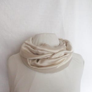 Peut inclure: Écharpe en satin crème drapée autour d'un buste de mannequin. L'écharpe est froncée et torsadée, créant un aspect texturé et superposé. Le tissu a un éclat subtil, reflétant la lumière. Le fond est blanc uni.