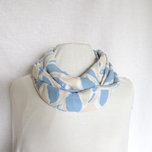 May include: A blue and cream-coloured infinity scarf on a mannequin. The scarf has a repeating pattern of blue shapes on a cream background. The fabric looks soft and lightweight, suitable for cooler weather.