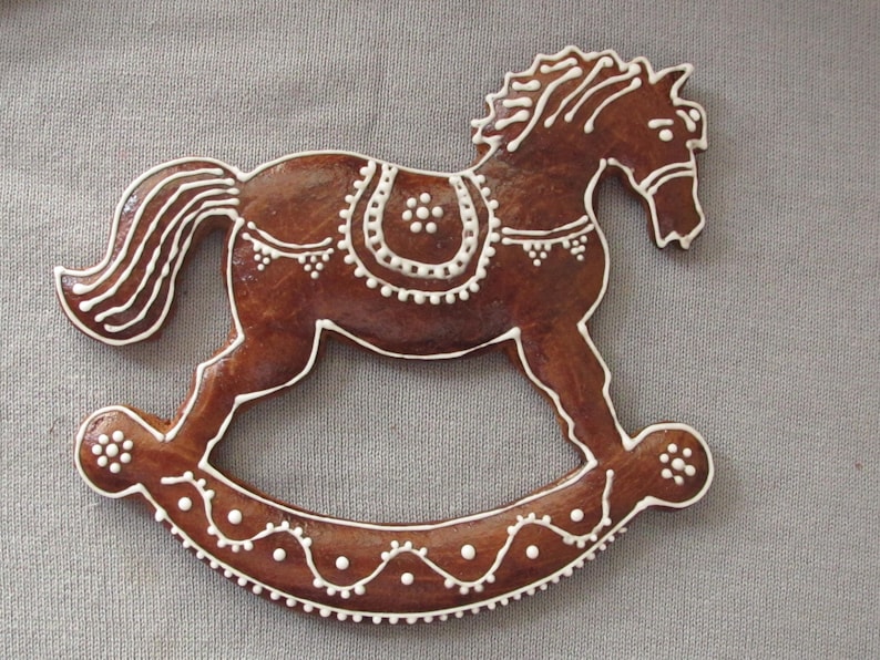 May include: A gingerbread rocking horse cookie decorated with white icing. The horse has a detailed mane and tail, and the rocking base is decorated with a wavy line and white dots.