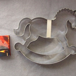 May include: A silver metal cookie cutter shaped like a rocking horse. The cutter is on a gray fabric background.