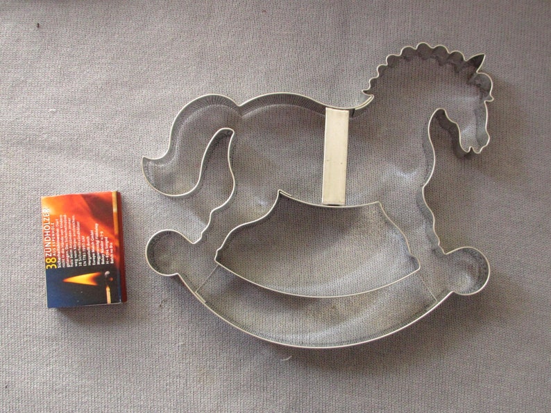 May include: A silver metal cookie cutter shaped like a rocking horse. The cutter is on a gray fabric background.