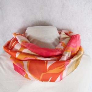 Könnte beinhalten: Ein rosa, orangefarbener und cremefarbener Seidenschal mit einem Blumenmuster. Der Schal ist gefaltet und auf einer weißen Oberfläche ausgestellt.