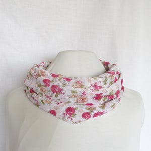 Op de afbeelding: Een witte infinity sjaal met een bloemenpatroon met roze en rode rozen, groene bladeren en bruine accenten. De sjaal is om de nek van een mannequin gedrapeerd en toont het ontwerp en de stijl. De sjaal is gemaakt van een lichte stof.