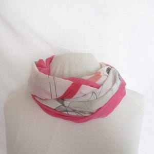 Può includere: Una sciarpa a cerchio rosa e bianca esposta su un manichino. La sciarpa presenta un bordo rosa acceso e un motivo di linee e forme rosa, grigie e nere. La sciarpa è realizzata in tessuto leggero.