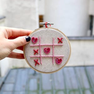 Peut inclure: Un cercle de broderie blanc avec un jeu de tic-tac-toe brodé en rose et rouge avec des cœurs au lieu des X et des O.