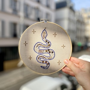 May include: Embroidered hoop art featuring a gold and blue snake with a beaded design on a beige background.