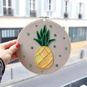 Könnte beinhalten: Ein runder Stickrahmen mit beigefarbenem Hintergrund, auf dem ein gelber Ananas mit grünen Blättern und kleinen grünen Blüten abgebildet ist.