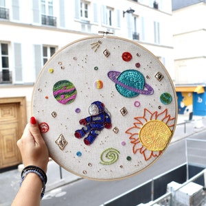 May include: Embroidered hoop art depicting a space scene with an astronaut, planets, stars, and a sun. The planets are embroidered with colorful beads and sequins.