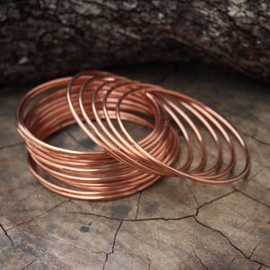 Könnte beinhalten: Eine Ansammlung kupferfarbener Armreifen, gestapelt und auf einer verwitterten Holzoberfläche angeordnet. Die dünnen, kreisförmigen Armbänder bestehen aus einem glänzenden, metallischen Material. Der Hintergrund zeigt eine verschwommene Holzstruktur.