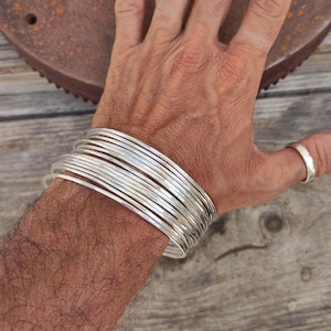 Puede incluir: Una pila de pulseras de plata en la muñeca de una persona. Las pulseras son finas y tienen una textura martillada.