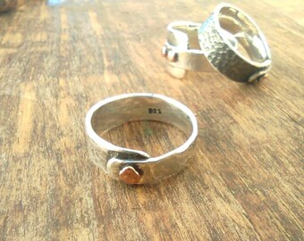 May include: Three silver rings with a hammered texture. The rings have a copper accent on the side. The rings are on a wooden surface.