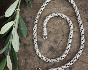Puede incluir: Un collar de cadena de plata con un diseño de cadena de caja. La cadena está sobre una superficie de madera con una planta verde en el fondo.