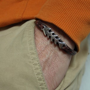 Puede incluir: Un brazalete de cuero marrón con un dije de hueso de pescado plateado. El brazalete se lleva en la muñeca.