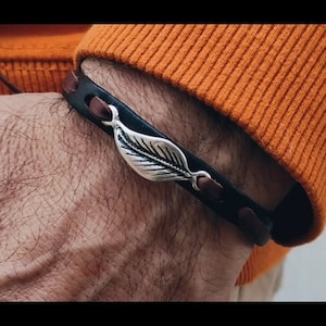 Può includere: Un braccialetto in pelle marrone con un ciondolo a forma di foglia d'argento. Il braccialetto è indossato al polso.