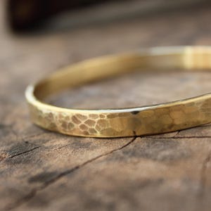 Puede incluir: Un brazalete dorado martillado. El brazalete tiene una superficie texturizada con una serie de pequeñas hendiduras irregulares. El brazalete tiene aproximadamente 6,35 cm de diámetro.