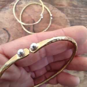 May include: A hammered brass bangle bracelet with two silver accents. The bracelet is open and has a hammered texture.
