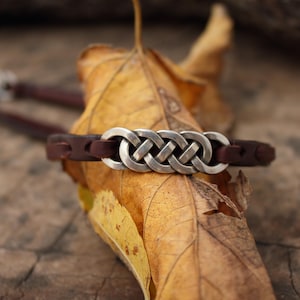 Puede incluir: Una pulsera de cuero marrón con un diseño de nudo celta plateado. La pulsera está sobre una hoja amarilla seca. El nudo plateado es el punto focal, con la correa de cuero a cada lado.