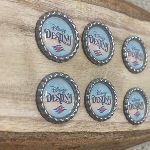 May include: Six bottle cap magnets with the text "Disney Destiny" on a wooden background. The magnets have a light blue background with the ship's logo. The bottle caps have a silver, textured edge.