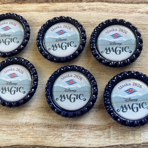 May include: Six navy blue bottle caps with a scalloped edge. Each cap features a scenic image with the text "Alaska 2026" and "Disney MAGIC". The caps are arranged on a wooden surface.
