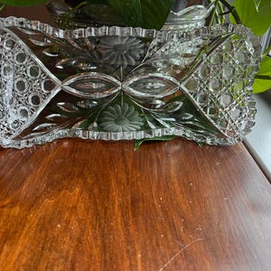 May include: A clear, rectangular glass dish with intricate cut glass patterns. The dish features a scalloped edge and floral and geometric designs. The dish is sitting on a wooden surface, with greenery in the background.
