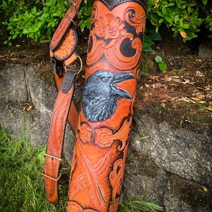 May include: A brown leather arrow quiver with intricate tooled designs, including a raven with black feathers and a floral pattern. The quiver has a shoulder strap and a flap with a fur lining.