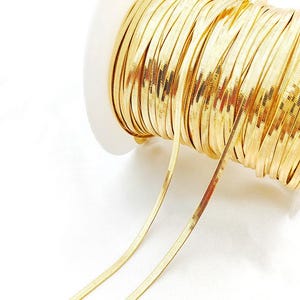 Könnte beinhalten: Eine Spule mit goldfarbener, flacher Fischgrätkette. Die Kette ist eng um eine weiße Kunststoffspule gewickelt. Zwei Stränge der Kette erstrecken sich von der Spule und zeigen das flache, strukturierte Design der Kette. Der Hintergrund ist weiß.