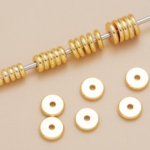 May include: A collection of small, round, gold-colored metal spacer beads with a smooth, reflective finish and a central hole. Several beads are threaded onto a thin silver-toned metal rod, while others are scattered loosely around the base.