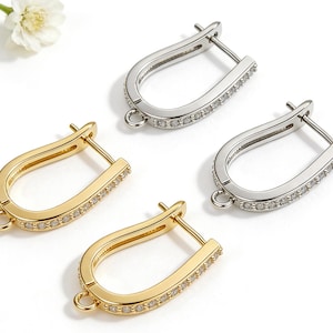 May include: Four U-shaped earrings, two gold-toned and two silver-toned, are displayed. Each earring is encrusted with small, clear gemstones along the outer curve. The earrings have a hinged closure and a small loop at the bottom for attaching charms.