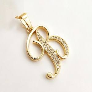 May include: Gold-colored pendant in the shape of the letter "B". The pendant is embellished with small, clear gemstones along the curves of the letter. The pendant has a loop at the top for a chain.