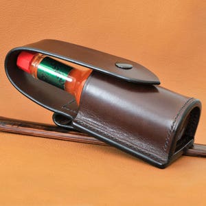 以下が含まれることがあります： 小さなホットソースのボトルを入れるための、スナップ留めの茶色の革製ホルスター。ボトルは赤と緑で、ラベルには「Sriracha」と書いてあります。