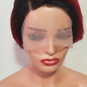 Op de afbeelding: Een kort, recht, synthetisch pruik met een ombre kleur van zwart naar bordeauxrood. De pruik heeft een lace front en is gestyled met een scheiding aan de zijkant.