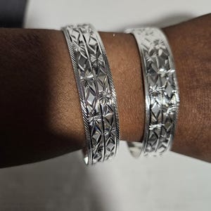 May include: Two silver bangles with detailed starburst and geometric patterns. The bracelets are worn on a dark-skinned arm, highlighting the intricate design and reflective surface. These bangles are a classic piece of jewellery.