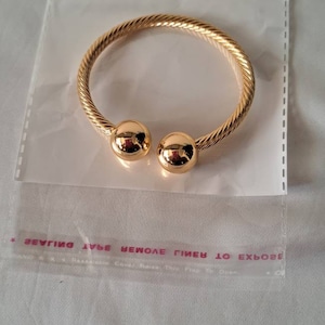 Può includere: Un braccialetto dorato con un design intrecciato e due accenti di sfera rotonda alle estremità. Il braccialetto è confezionato in un sacchetto di plastica trasparente con un'intestazione rossa che dice "LS Jewelry".
