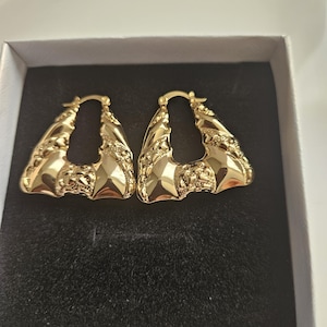 May include: A pair of gold-colored, textured hoop earrings. The earrings are triangular in shape with a detailed, ornate design. They are displayed inside a white box with a black interior.
