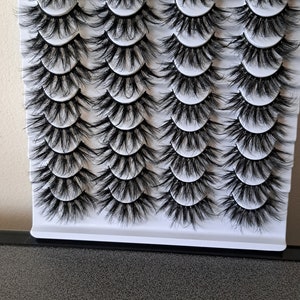 Op de afbeelding: Een witte plastic bak met 25 individuele zwarte valse wimpers. De wimpers zijn in rijen gerangschikt en klaar om te worden aangebracht.