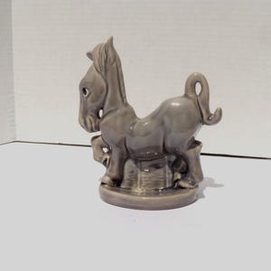 May include: A glossy, light gray ceramic figurine of a horse standing on a circular base. The horse is depicted in a stylized pose with its head turned to the side, a raised front leg, and a curved tail. The smooth, reflective surface is uniform.