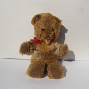 May include: A brown teddy bear with a red bow tie and a tongue sticking out. The bear has a stitched nose and brown button eyes.