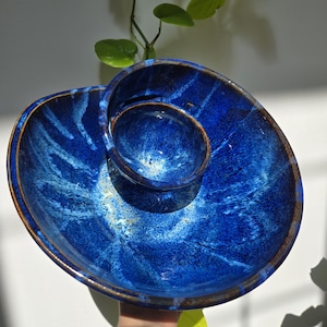 Può includere: Un piatto da portata in ceramica fatto a mano con una glassa blu intenso e una ciotola per salse integrata. Il piatto ha una forma organica unica con un bordo marrone. La glassa presenta striature bianche e azzurre, creando un effetto marmorizzato. Perfetto per servire snack o antipasti.
