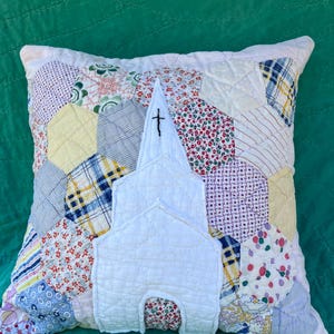 Puede incluir: Un cojín cuadrado con un diseño de patchwork hecho de piezas de tela hexagonales multicolores. Centrado en el cojín hay un aplique de tela blanca con forma de edificio de iglesia con un campanario y una pequeña cruz negra bordada.