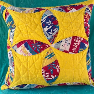 May include: A square quilted throw pillow featuring a bright yellow fabric overlay with a central four-petal floral cutout design. The cutout reveals a multi-colored patterned fabric underneath with red, blue, and white floral and leaf motifs.