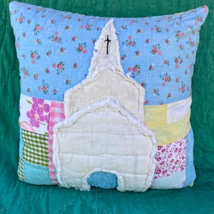 May include: A square quilted throw pillow featuring a patchwork design of light blue floral, pink floral, yellow, and green gingham fabrics. Centered on the front is a white fabric applique shaped like a church with a black embroidered cross.