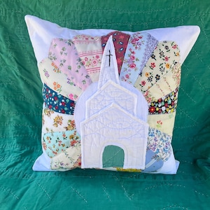 May include: A square throw pillow featuring a central white fabric applique of a church with a steeple and a green door. The background consists of a fan-shaped pattern made from various floral-printed fabric segments in pink, blue, and yellow.