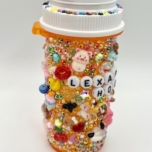 Puede incluir: Un frasco de pastillas naranja decorado con brillantes de colores y varios abalorios, incluyendo un cerdo, un beso y un oso. La tapa blanca del frasco también está adornada con brillantes. El nombre "LEXA HO" está escrito con cuentas de letras blancas en el frasco.