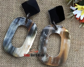Può includere: Un paio di orecchini in corno con un perno quadrato nero e una grande forma rettangolare aperta. Gli orecchini presentano una palette di colori naturali di crema, marrone e grigio. Gli orecchini sono presentati su una superficie beige strutturata.