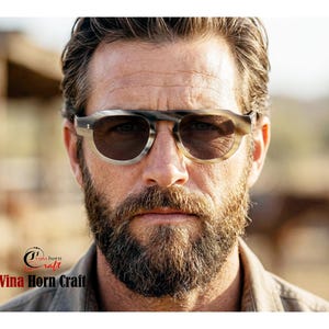 May include: A person wearing sunglasses with a light brown and grey frame and dark lenses. The sunglasses have a rounded shape. The person has a full beard and is wearing a light-coloured shirt. The text "Vina Horn Craft" is visible.