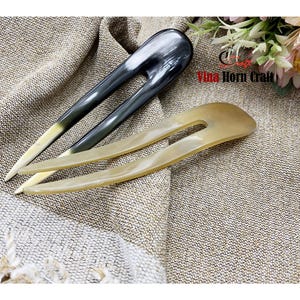 Puede incluir: Dos horquillas para el cabello de cuerno. Una es gris oscuro con punta blanca, la otra es de color beige claro. Los accesorios para el cabello est&aacute;n sobre una tela texturizada de color beige. La imagen est&aacute; bien iluminada, mostrando las variaciones naturales del material de cuerno.