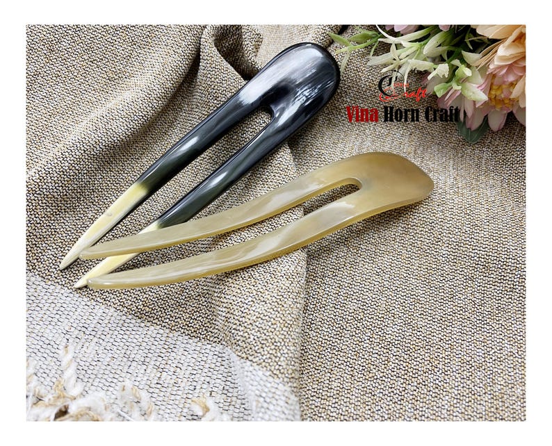 Puede incluir: Dos horquillas para el cabello de cuerno. Una es gris oscuro con punta blanca, la otra es de color beige claro. Los accesorios para el cabello est&aacute;n sobre una tela texturizada de color beige. La imagen est&aacute; bien iluminada, mostrando las variaciones naturales del material de cuerno.