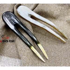 Puede incluir: Dos horquillas para el cabello hechas de cuerno. Una es gris oscuro y la otra blanquecina. Ambas tienen un acabado liso y pulido y un dise&ntilde;o de dos puntas. Las puntas de las horquillas son de un color m&aacute;s claro, creando un efecto degradado. Las horquillas se exhiben sobre una tela beige texturizada.